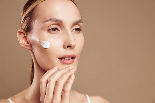 Young woman with clear skin applying white face cream on cheek, close-up portrait on beige background, concept of skincare, beauty, wellness, and daily self-care routine with natural light.