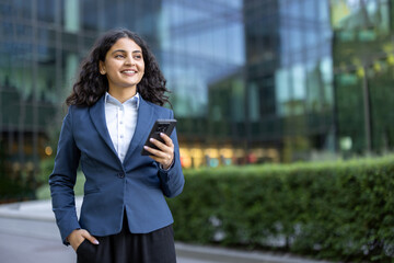 Young professional businesswoman smiling and holding a smartphone. Looking away in a modern urban...