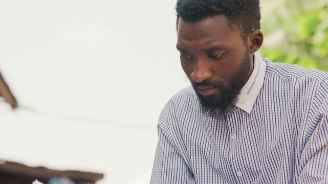 Black man focused eyes down outdoors, patterned shirt and beard in calm tropical light determined expression suggests planning or study, quiet entrepreneurial or student energy with reflective mood