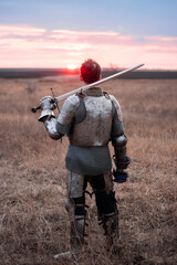 A knight in cuirass and chainmail, holding a sword and a bloodied face, stands with his back to the sunset in a field. A medieval warrior after a battle. © Михаил Гута