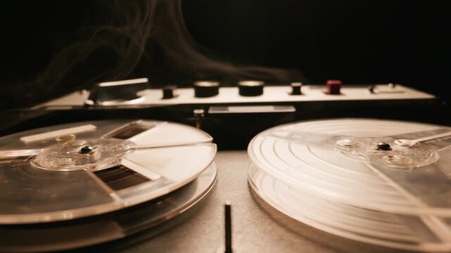 Vintage reel to reel tape recorder plays as cinematic smoke drifts across spinning spools. Warm dramatic lighting highlights the mechanical details of the retro analog audio machine.