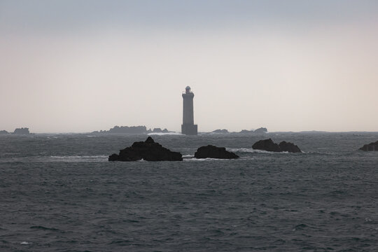 View of the Phare de la Jument lighthouse standing amidst crashing waves and dark jagged rocks under a moody overcast sky in Ushant, Brittany, France.