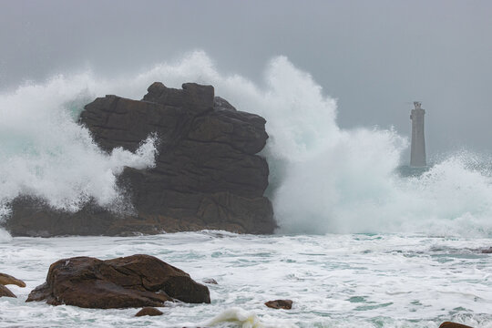 View of powerful waves crashing against dark coastal rocks with the Nividic Lighthouse visible in the misty background in Ushant, Brittany, France.
