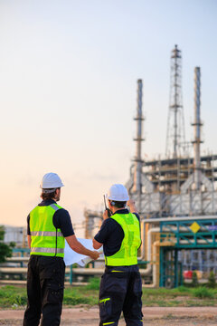 Two professional male engineers wearing safety gear standing outdoors at dusk, discussing blueprints and project details in front of a large oil refinery and petrochemical manufacturing facility.