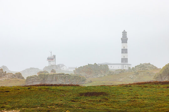 View of Phare du Creac'h lighthouse and signal station among rocky outcrops and green fields under a thick fog Ushant, Brittany, France.