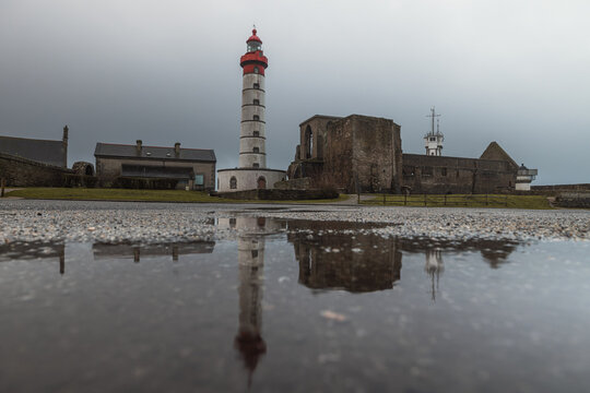 View of the Saint-Mathieu Lighthouse and abbey ruins reflected in a puddle under a cloudy sky at Pointe Saint-Mathieu, Plougonvelin, Brittany, France.
