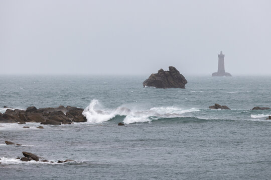 View of Phare du Four lighthouse with crashing waves against rocky shores under a misty sky in Porspoder, Brittany, France.