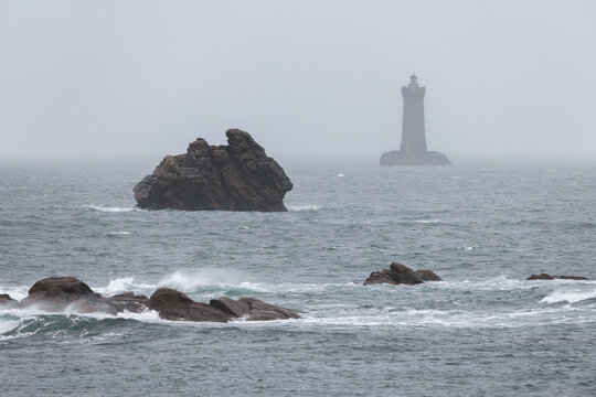 View of Phare du Four lighthouse standing amidst rough sea and rocky outcrops under a foggy sky in Porspoder, Brittany, France.