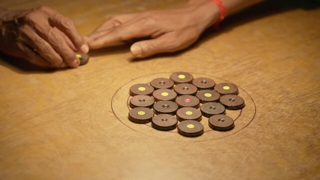 Multiple player hands arranging carrom coins in center circle on wooden board, preparing setup before play, indoor scene with warm light showing traditional game