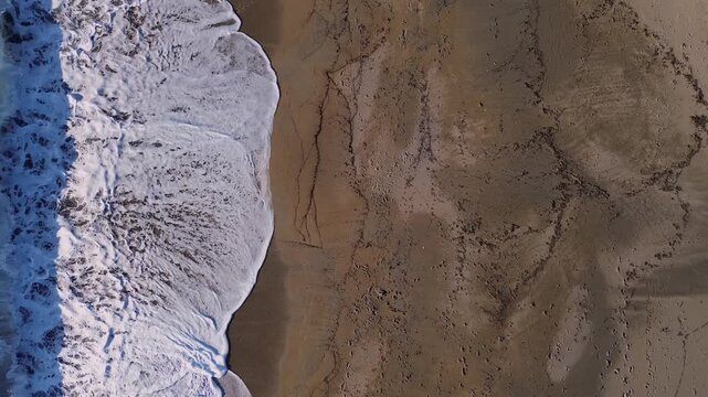 Cinematic top-down drone shot of the vast Monolithi beach. High-angle view of sea waves washing onto the sandy shore, revealing footprints and organic textures on the wet sand. Stunning Ionian sea.