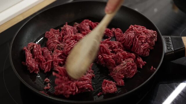Frozen raw minced beef in frying pan with wooden spoon preparing for cooking stage.