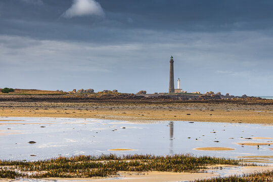 View of Ile Vierge lighthouse standing tall on a rocky island across a sandy beach with water reflections under a cloudy sky Plouguerneau, Bretagne, France.