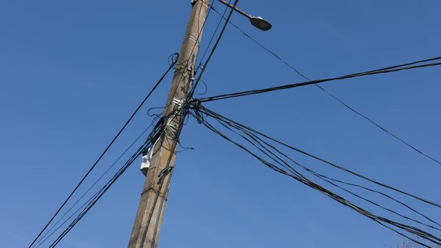 Concrete utility pole holds a chaotic tangle of black electrical and communication cables under a bright clear blue sky. The scene illustrates an urban infrastructure overload concept.