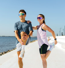 Athletic man and woman stretch legs by the waterfront promenade on a bright sunny day, preparing for exercise