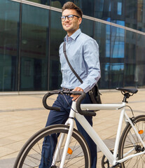 Smiling professional man in glasses walks a white bicycle outdoors near a modern glass office building during daytime