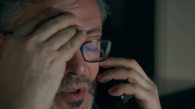 Close-up of face of 50-year-old Caucasian man talking to wife or girlfriend on mobile phone, while working late at night in dark office, blue computer screen reflecting on glasses