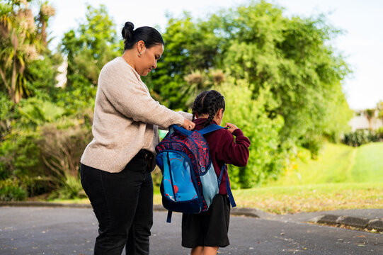 Niuean mum helping son put on his backpack while standing on paved footpath