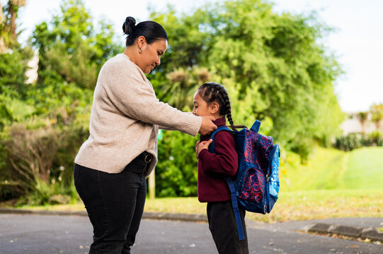 Niuean mum helping son put on his backpack while standing on paved footpath