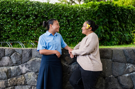 Niuean mum and teenage daughter having a conversation while leaning against stone ledge outdoors