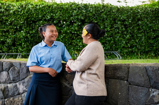 Niuean mum and teenage daughter having a conversation while leaning against stone ledge outdoors