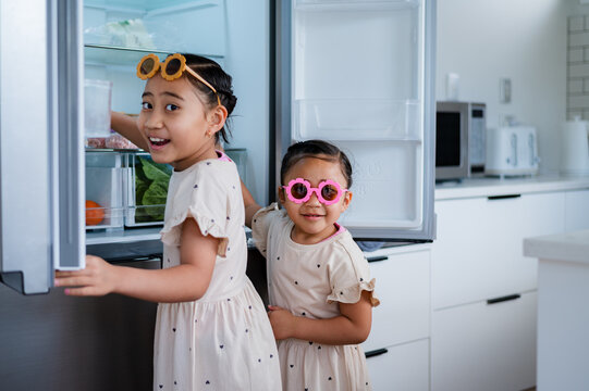 Two young girls wearing flower sunglasses opening the fridge looking for a snack