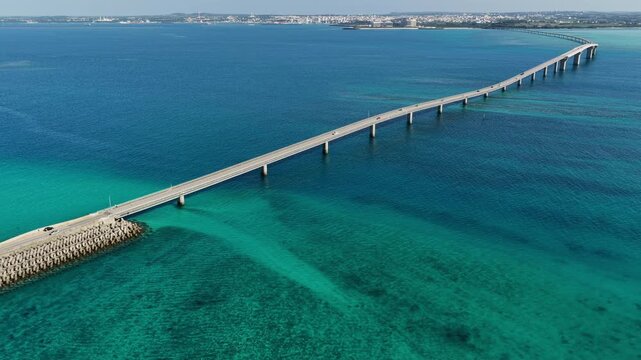 Aerial bird's eye view of Irabu Bridge in Miyako Island, Okinawa. Diagonal composition over emerald blue sea with a boat approaching the bridge.