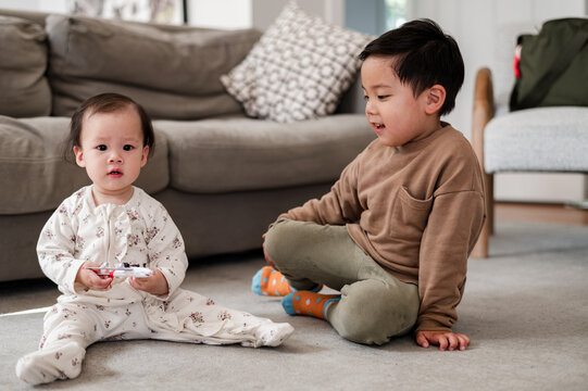 Asian toddler boy looking at baby sister both seated on carpeted floor in the living room