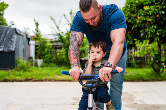 Father guiding toddler son on riding his bicycle on backyards paved surface