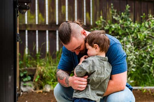 Father with tattooed arm hugging toddler son while crouching in the backyard