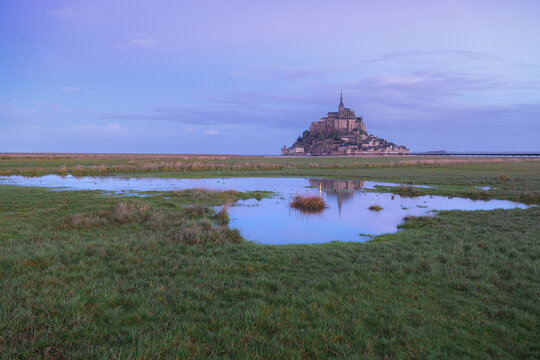 View of the historic Mont Saint-Michel abbey rising above green marshland with water reflections under a twilight sky in Mont Saint-Michel, Normandy, France.