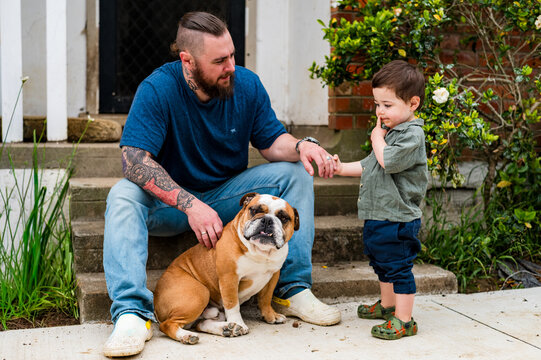 Father sitting on a step holding toddler son and petting dog seated on the ground