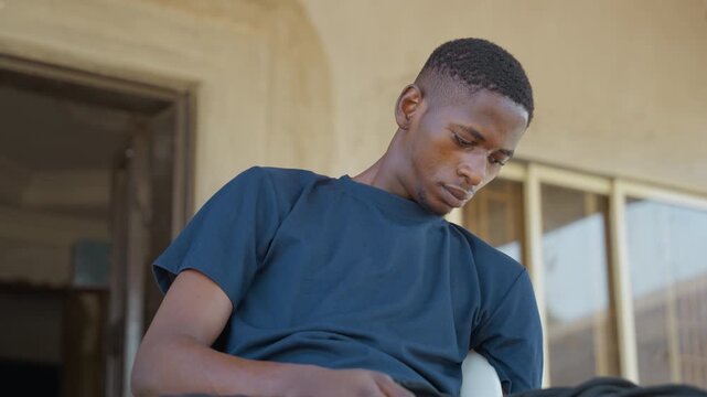 Young man on porch wearing black shirt, sitting on house steps after chores, looking down in quiet reflection, warm sunlight on doorway, casual tshirt and relaxed posture conveying fatigue