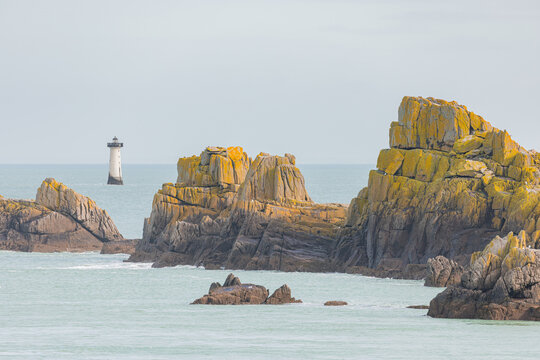 View of the jagged rocks at Pointe du Grouin with a lighthouse in the distance across the turquoise sea in Cancale, Bretagne, France.