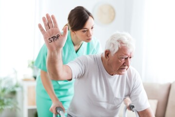 Obraz premium Senior Man with Walker Protests Unwanted Help from Female Nurse