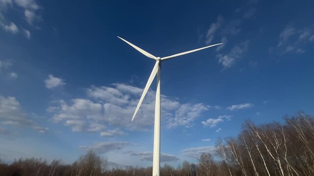 Wind turbine spins against clear blue sky. Clouds drift gently around its white blades. No people appear in this scene. Ideal for renewable energy or environmental visuals