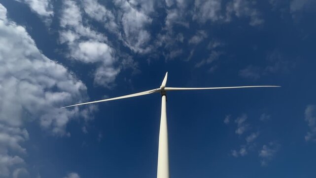 Wind turbine spins against clear blue sky. Clouds drift gently around its white blades. No people appear in this scene. Ideal for renewable energy or environmental visuals