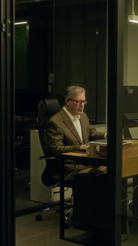 Wide vertical shot of stressed middle-aged Caucasian man working on report or project at desk in office at night, staring at computer screen, taking off glasses, massaging face and drinking coffee