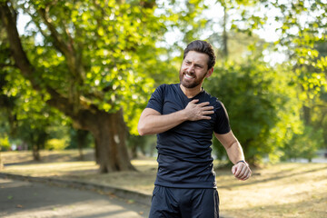 A young male athlete, twisted in pain, stands in the park and holds his hand on his shoulder © Tetiana