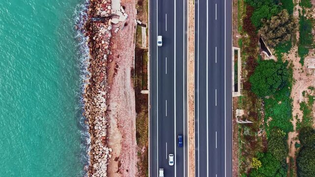 Cars travel along the highway beside the ocean. The road is straight and runs parallel to the water. The coastline has rocks and greenery.