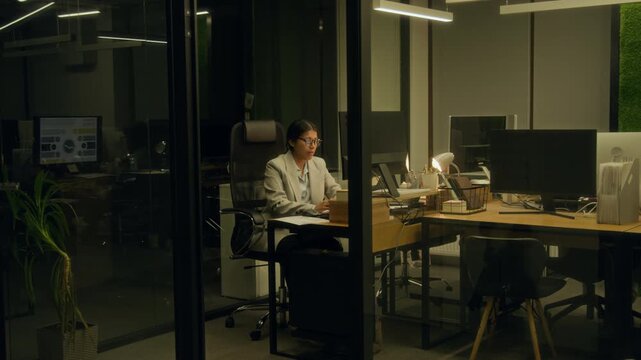 Wide shot of middle-aged Hispanic female accountant working on financial report late at night in dark office, declining personal phone call, taking off glasses and rubbing sore eyes with exhaustion