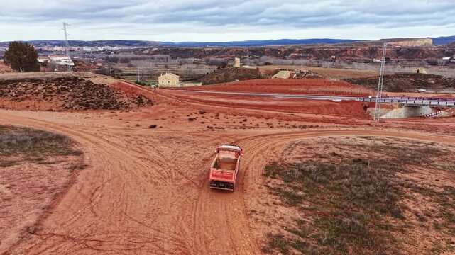 A truck travels down a dirt road in a rural setting with hills and distant buildings. The ground is dry with patches of grass. The sky is cloudy, adding a gray tone to the scene.