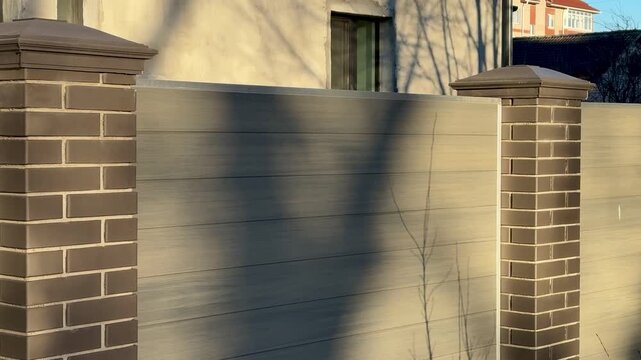 Sunlit brick column by cottage fence, warm golden hour light casts long tree shadows across weathered wooden panels, capstone.