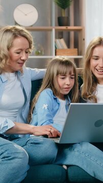 Attractive carefree joyful friendly loving mom and her younger and eldest daughters sitting on comfortable sofa in cozy living-room during video conversation on laptop