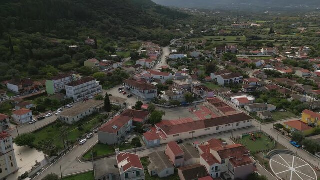 An aerial view captures a tranquil Greek village nestled amidst verdant hills, showcasing a charming mix of traditional and modern architecture, streets, and a schoolyard.