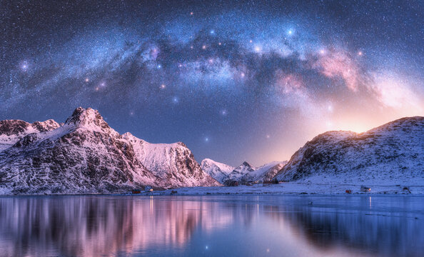 Milky Way arch above sea coast and snow covered mountains in winter at night. Lofoten Islands, Norway. Arctic landscape with blue starry sky, arched milky way reflected in water, snowy rocks. Space
