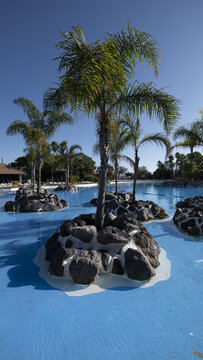 View of a large blue swimming pool with artificial rock islands and palm trees under a clear sky in Ceuta, Ceuta, Spain.