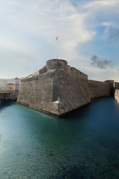 View of the historic Royal Walls and the San Felipe Moat with clear turquoise water and a flag flying under a bright sky in Ceuta, Ceuta, Spain.