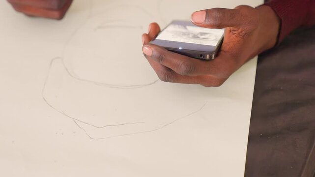 Black hand holds phone over paper, darkskinned hands alternate between thumb scrolling reference and pencil marking outline on white sheet, closeup of grip and screen glow, intimate creative moment