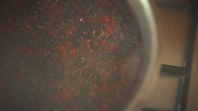 In a home kitchen, a metal bowl on the stove simmers a mix of berries, beginning the jam making process. The berries bubble as sweet homemade berry jam takes shape, filling the air with fruity aroma