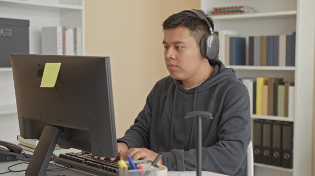 Man with headphones typing on keyboard at a computer desk in a building office, hands on keyboard and neutral expression; concentration.